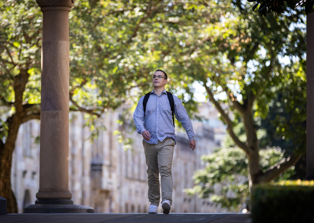 Image of a young man with very short hair, glasses, white jeans and a blue long-sleeve shirt. He is walking through the sandstone columns outside at the Great Court at UQ