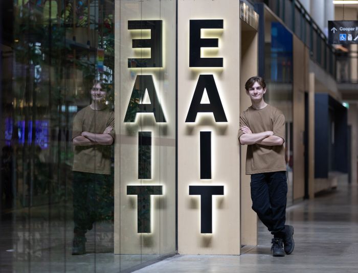 Image of a young man with short hair and brown shirt. He is standing next to a sign that says 'EAIT'.