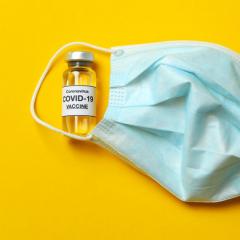 Vial of Covid-19 vaccine and blue mask on yellow background