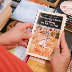 An old book held by two hands over a stack of other old novels.