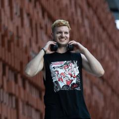A blond man in a black and red band tee and black headphones smiles towards the camera.