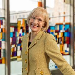 A woman in a beige suit smiles towards the camera. In the background is a colourful stained glass window.