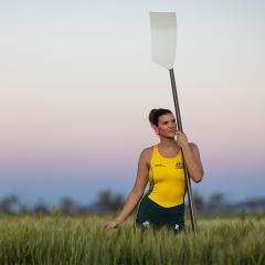 An image of UQ agribusiness student and Australian rowing representative standing in a paddock of wheat rows at UQ's Gatton campus. She is wearing her Australian rowing zoot suit and is leaning on an oar as the sun sets.