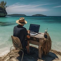 A digital nomad working on a laptop while sitting on a beautiful beach with turquoise water in the background.