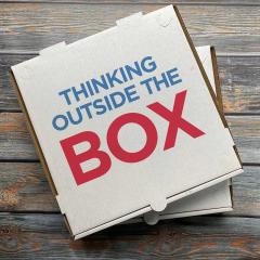 An image of a pizza box resting on a wooden table. On the lid of the box it reads, 'Think outside the box'.