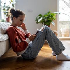 Teenage girl sitting on floor near bed using smartphone at home, scrolling social media.