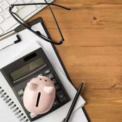 A top view image of a notepad, calculator, piggy bank and reading glasses on a dark wooden tabletop.