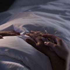 An image of the hands of a terminally ill on a hospital bed with a loved placing their hand on the patient's.