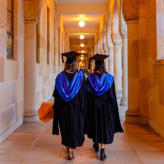 An image of two graduates wearing cap and gowns, walking through the cloisters of UQ's Great Court. walking