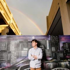 An image of UQ Doctor of Medicine student and The VacSeen Project co-director Jeremy Hunt standing in front of a cityscape mural in Brisbane. A rainbow is beaming through stormy skies in the background..