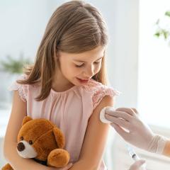 An image of a young girl holding a teddy bear while receiving an injection.