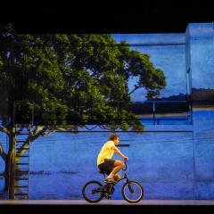 A young man wearing a yellow t-shirt rides a bicycle on a theatre stage.