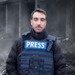 A man in a press flak jacket standing on a road surrounded by rubble.