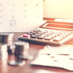 A photo of a calculator with stacks of coins in the foreground, and a calendar in the background