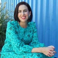 Sarah Holland-Batt, a woman with shoulder-length dark hair, smiling and sitting in front of a blue shipping container while wearing a bright blue dress and blue earrings