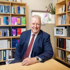 Professor Matthew Sanders, an older man, sitting and smiling at a desk in an office in front of two full bookshelves and a small filing cabinet