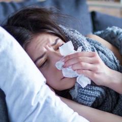 A woman lying underneath a blanket on a couch with a pillow under her head and blowing her nose.