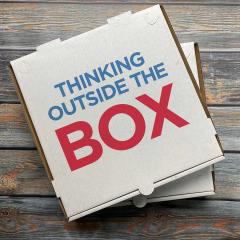 An image of a pizza box resting on a wooden table. On the lid of the box it reads, 'Think outside the box'.