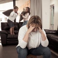 An overwhelmed mother struggling with kids having pillow fights and jumping on couch.