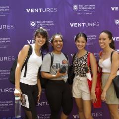 Four women smiling at the camera, they are standing in front of a purple background.