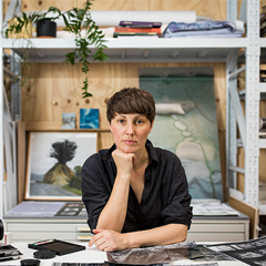 Image of a woman sitting at a desk with her head resting on her chin, she is looking into the camera